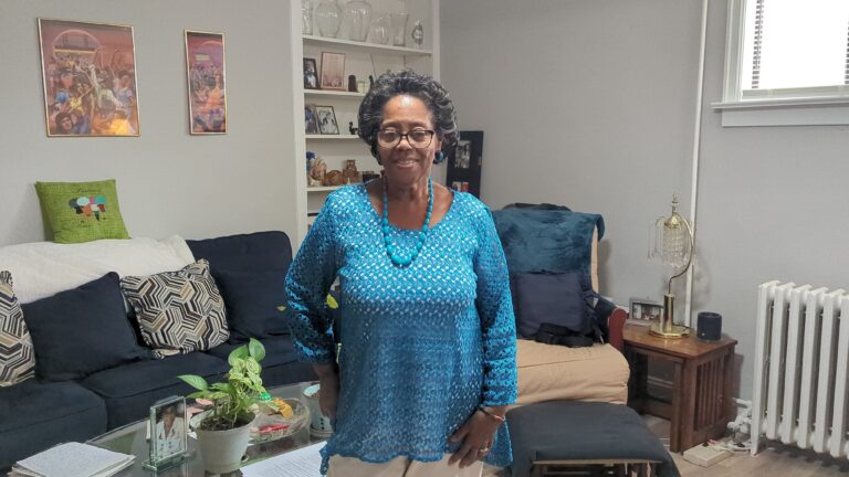Monnie Early in a bright blue shirt smiling in her newly renovated living room at 49-53 Missouri Ave