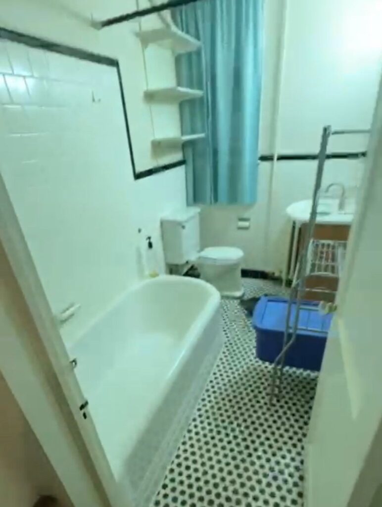 Pre-renovation bathroom at 49-53 Missouri features a tub, outdated black and white floor tiles, dingy white tile on the walls, with an aqua blue curtain covering the window.