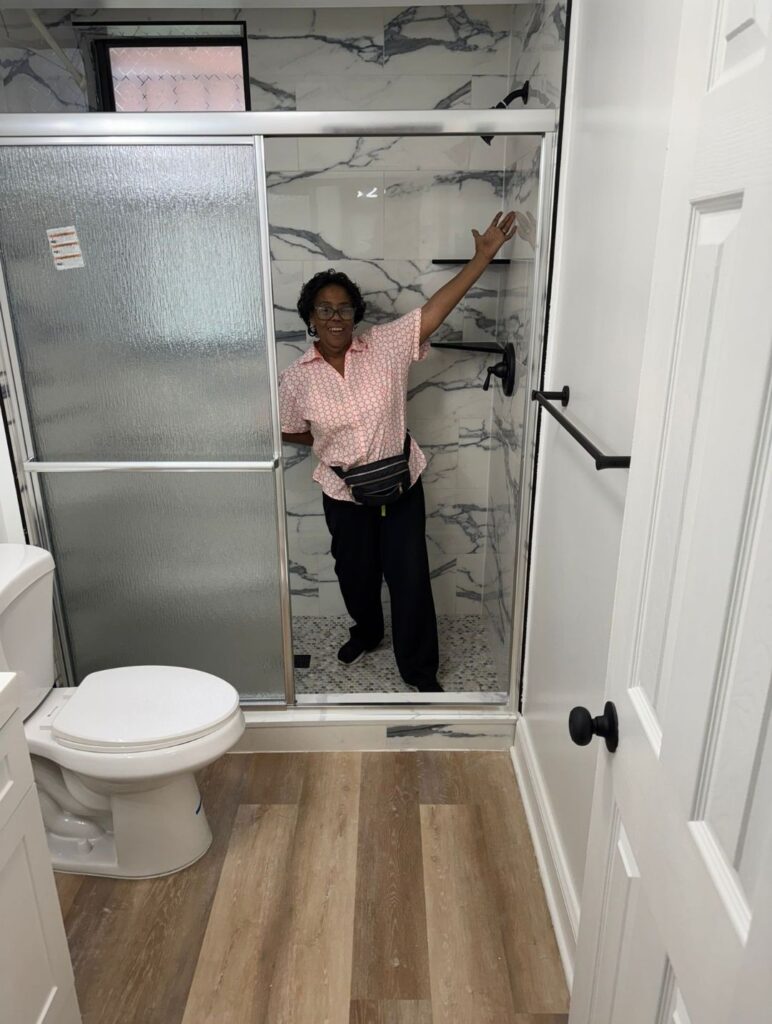 Monnie Gordon, dressed in a red and white blouse and black jeans is smiling with her arms open in her newly renovated walk in shower that features marble tile and sleek, black handles and a tile rack.