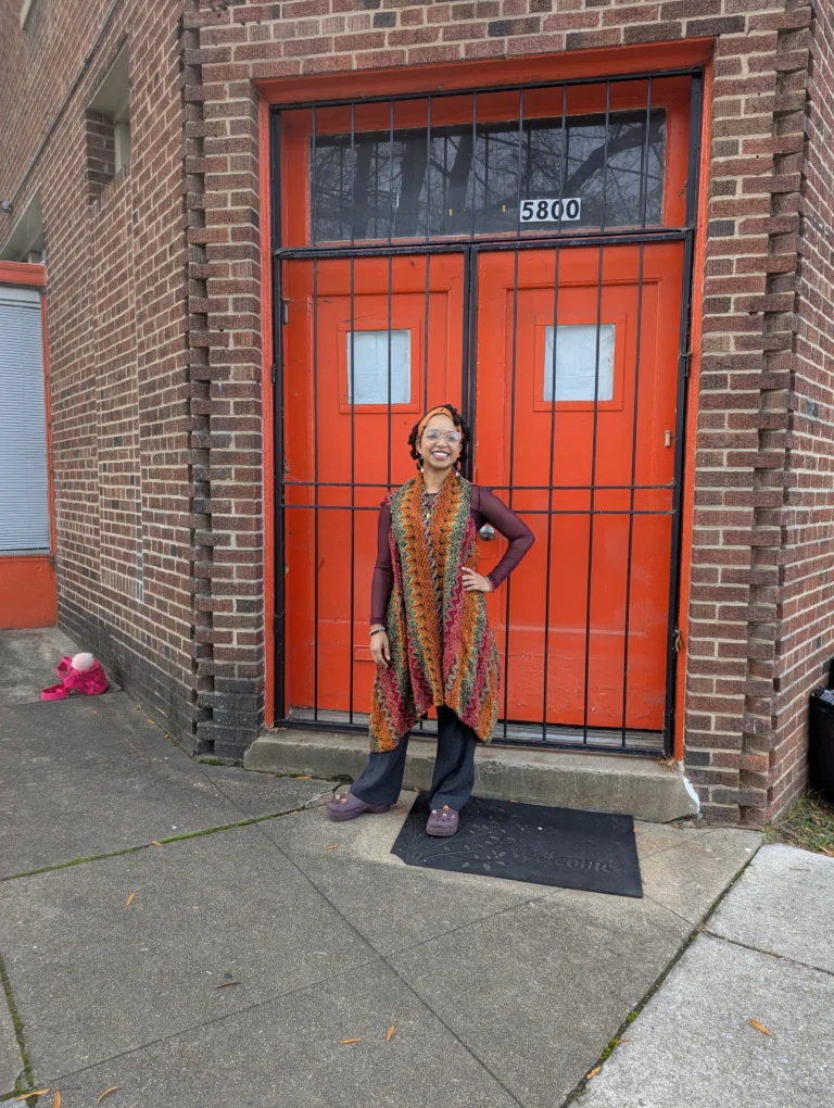 Chelsea Harrison strikes a pose in a burnt orange, magenta, and olive green crochet sweater in front of a pair of bright orange doors in a brick facade that lead to the entrance of 11 Nicholson's community room