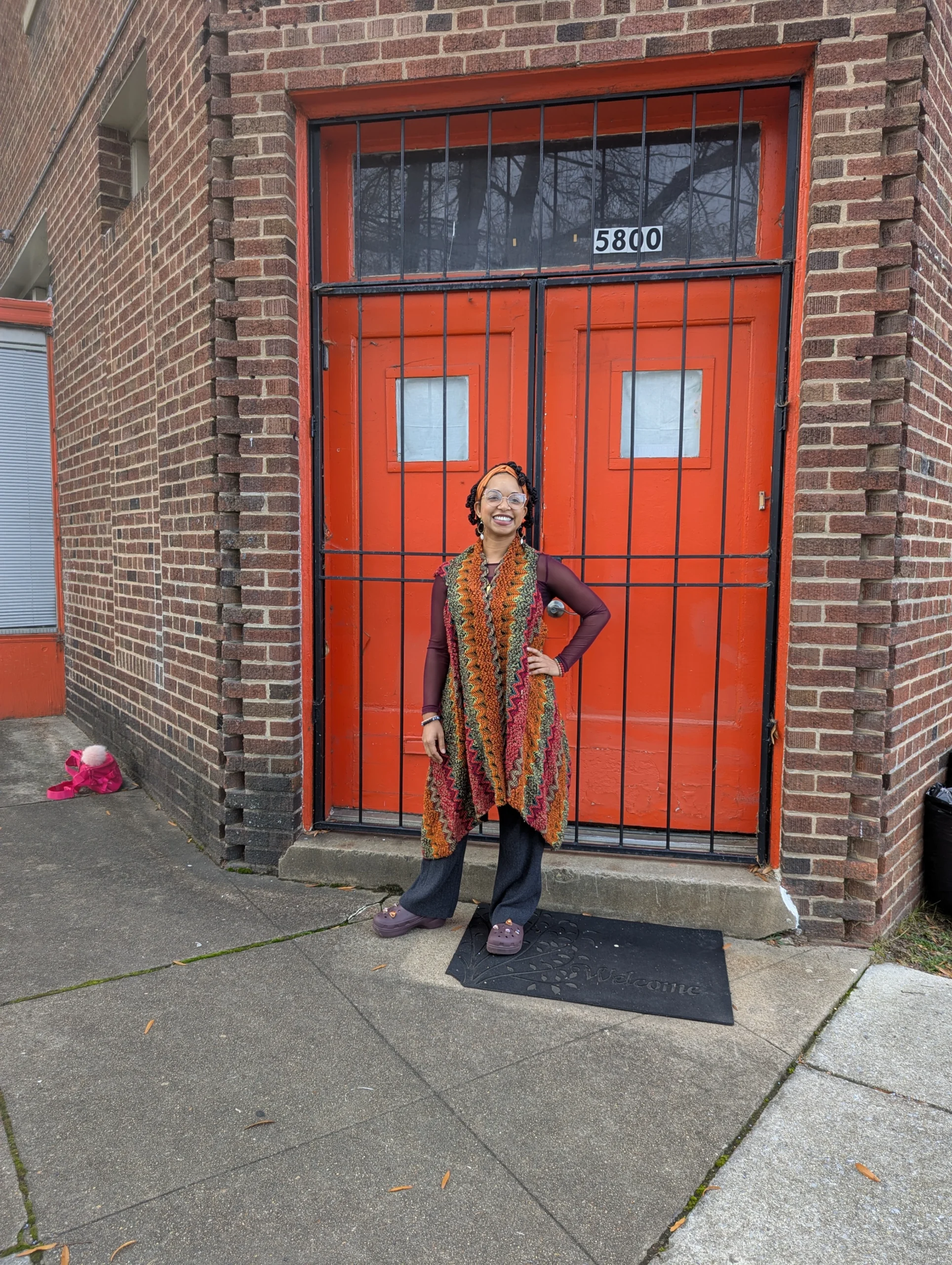 Chelsea Harrison strikes a pose in a burnt orange, magenta, and olive green crochet sweater in front of a pair of bright orange doors in a brick facade that lead to the entrance of 11 Nicholson's community room