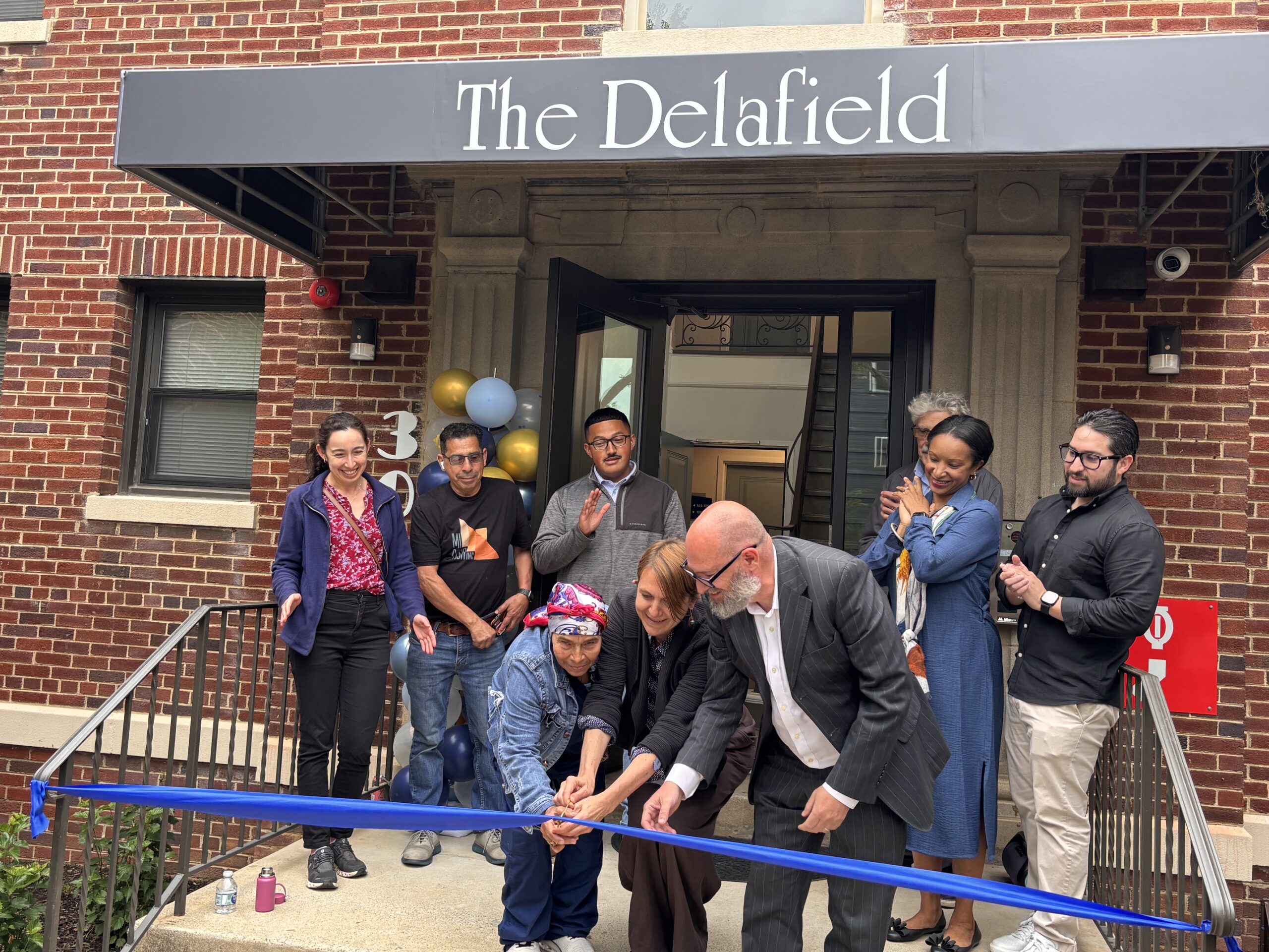 Resident and Co-Executive Directors smile as they cut the ribbon in front of the newly renovated 301 Delafield apartment building